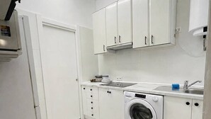 Een kleine keuken met witte kastjes, een wasmachine, een spoelbak en een tafel.