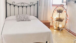 Een bed met een metalen frame, een witte spreideken, een kussen met een patroon, een klein houten nachtkastje, een raam met witte gordijnen en een decoratieve plantenstandaard.