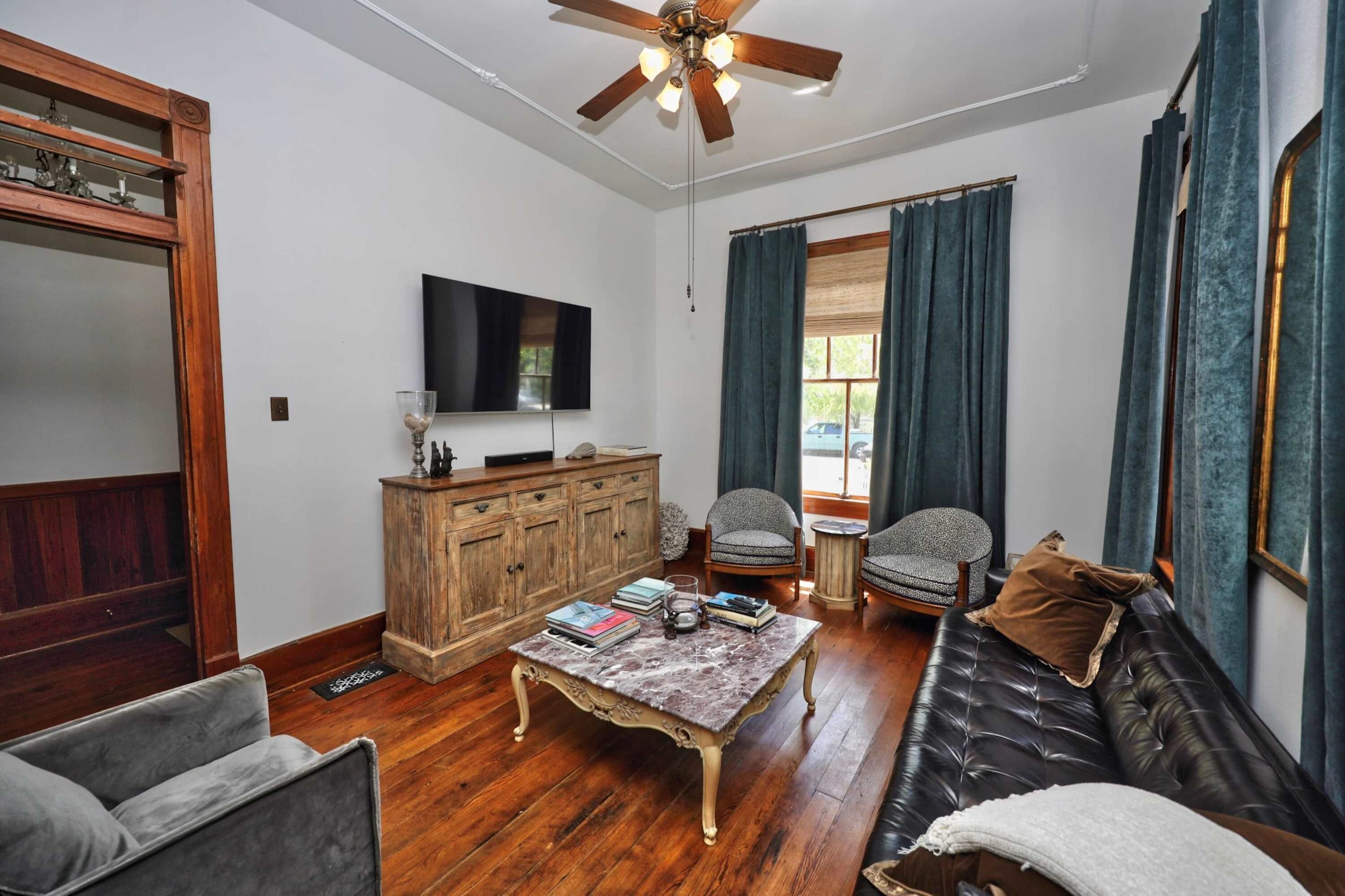A living room with a wooden floor, a ceiling fan, a television, a sofa, two armchairs, a coffee table, and a cabinet.