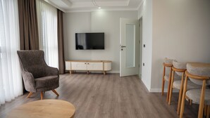 A modern living room with a large flat-screen TV, a brown armchair, a wooden coffee table, and a dining area with three bar stools. - Santana Suit (Arsin)