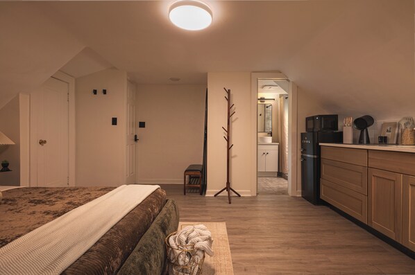 Ein Schlafzimmer mit einem Bett, einem Nachttisch, einer Lampe, einem Kleiderhaken und Blick ins Badezimmer.