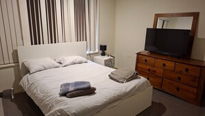 Un letto rifatto con cuscini, una lampada da comodino, un televisore su una cassettiera in legno e una finestra con persiane.