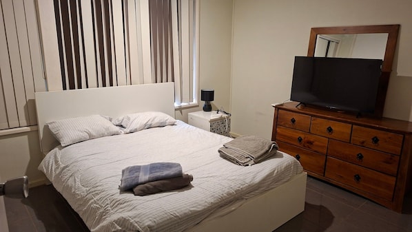 A neatly made bed with pillows, a bedside lamp, a television on a wooden dresser, and a window with blinds.