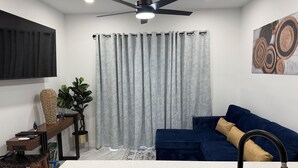 A modern living room with a dark blue sofa, a white countertop with a sink, a ceiling fan, and a grey curtain.