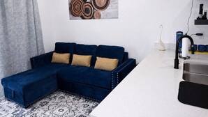 A modern living room with a blue sectional sofa, a patterned rug, a kitchen area with a sink and countertop, and a wall-mounted artwork.
