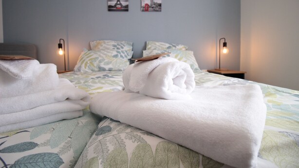 A neatly made bed with folded towels, a patterned bedspread, and two bedside lamps. - Les Loges de Véro RDC - 5 min from Verdun, private parking, motorcycle garage, air conditioning. (Thierville-sur-Meuse)