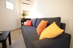 Un salon avec un canapé gris foncé, deux coussins orange et un coussin jaune, une table basse noire et une petite plante posée sur une table d’appoint.