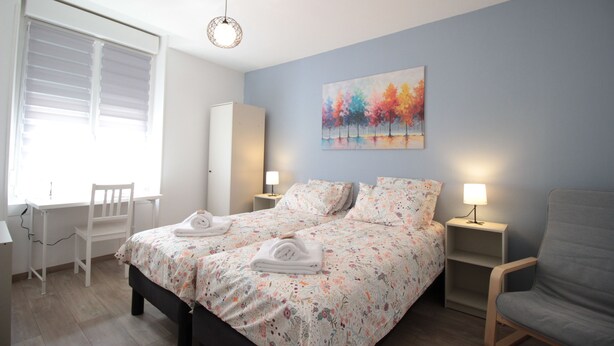 A bedroom with a bed, two bedside tables, a chair, a desk, and a window with blinds.