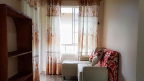 A room with a sofa, a wooden shelf, a window with curtains, and a tiled floor.