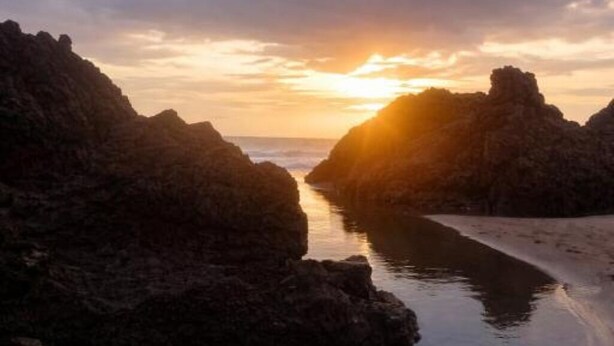 A coastal landscape with rocky formations and a calm body of water reflecting the sunset.