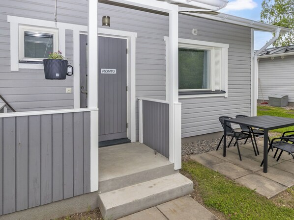A small, single-story house with a gray exterior, a front door, a window, and a patio area with a table and chairs.