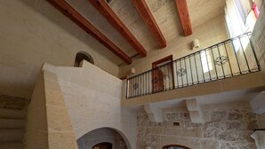 Interior de pedra com escadaria de madeira, varanda com grades de ferro forjado e parede de pedra com janela.