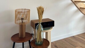 Una stanza con soffitto in listelli di legno, due quadri appesi al muro, una lampada e un vaso con steli di grano su uno sgabello.