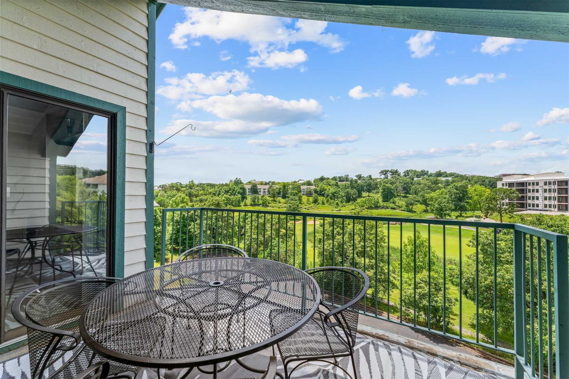A balcony with a table and chairs, overlooking a green landscape with trees and buildings.