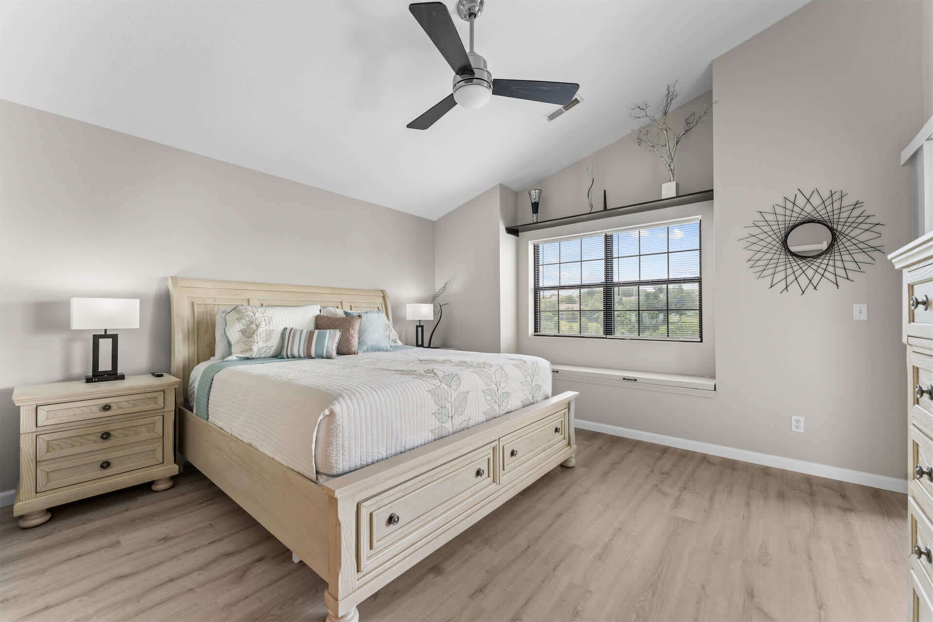 A bedroom with a bed, nightstands, a ceiling fan, a window with blinds, and a wall-mounted decorative piece.
