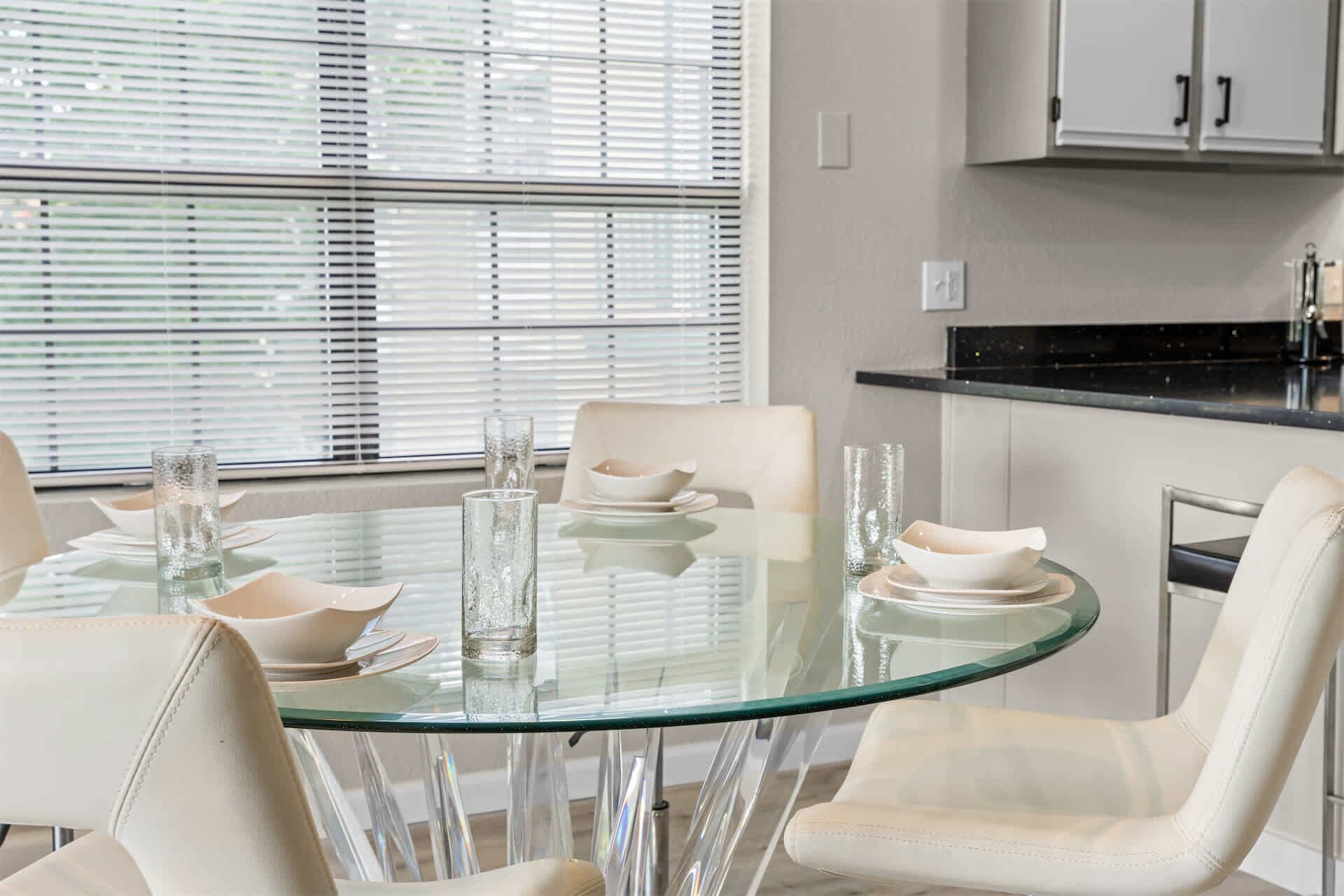 A dining area with a round glass table, white chairs, and a window with blinds.