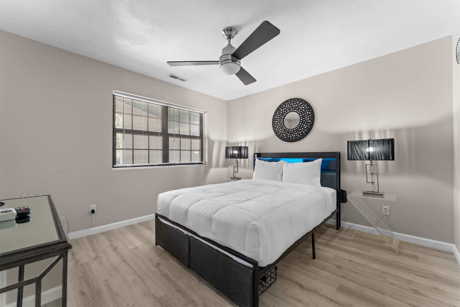 A bedroom with a bed, ceiling fan, bedside lamps, a round mirror, and a window with blinds.