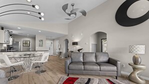 Een moderne woonkamer met een grijze leren bank, een eethoek met een glazen tafel, en een keuken met witte kasten.