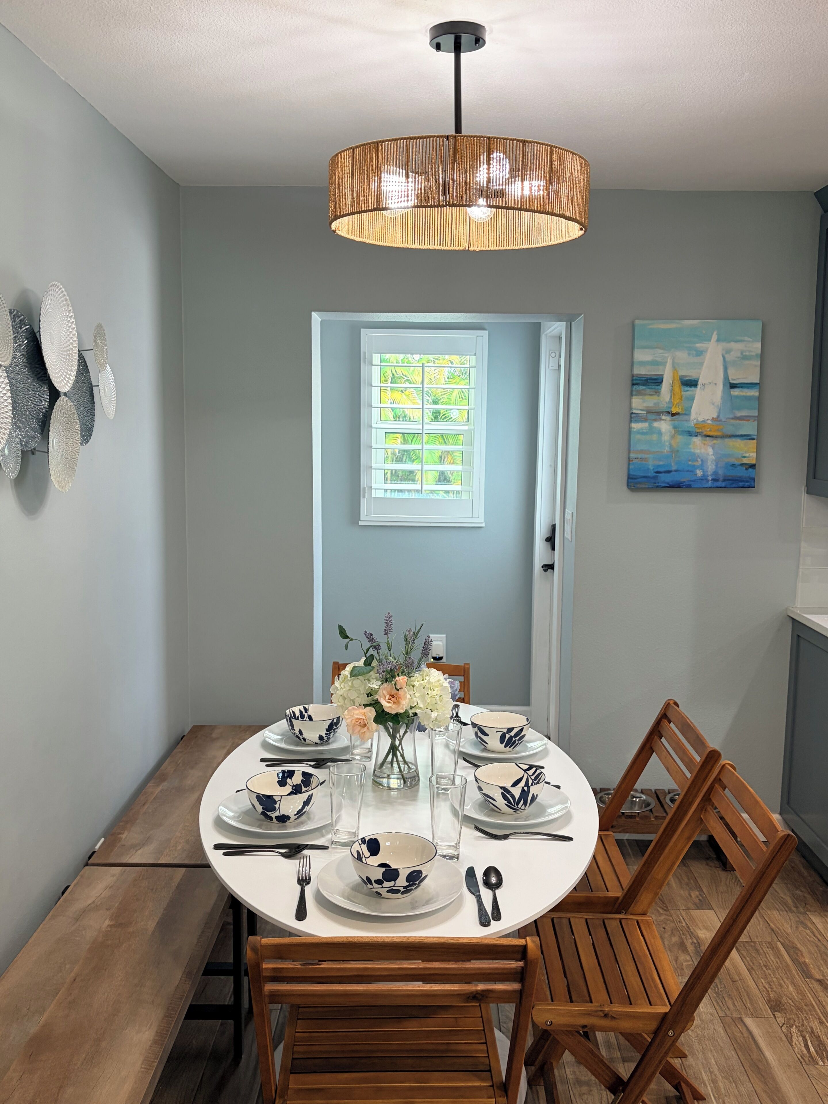 A dining area with a round table set for four, a vase of flowers, and a painting on the wall.