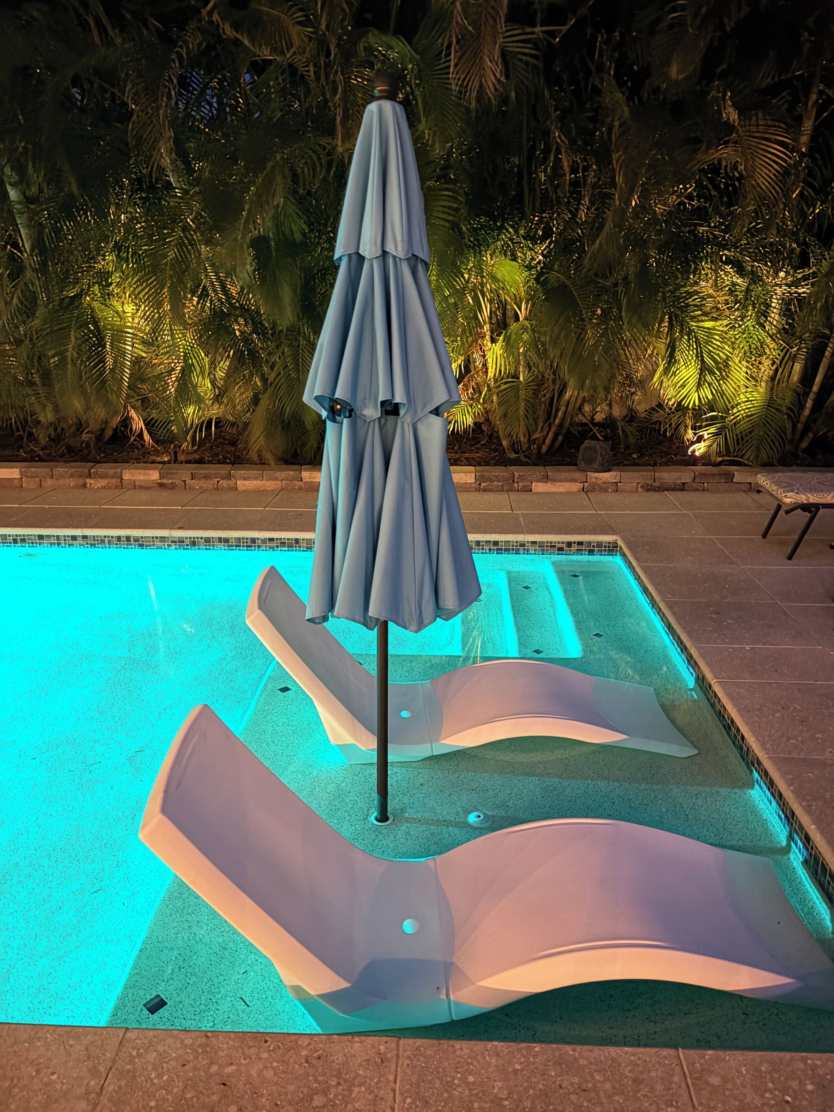 Una piscina con borde iluminado en azul, sillas de descanso y una sombrilla.