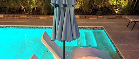 Una piscina con borde iluminado en azul, sillas de descanso y una sombrilla.