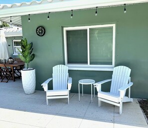 Eine Terrasse mit zwei weißen Adirondack-Stühlen, einem kleinen runden Tisch, einem Blumentopf und einer an der Wand befestigten Uhr.