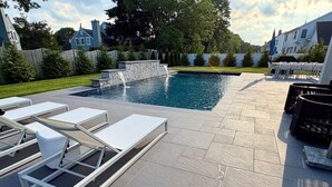 A swimming pool with a waterfall feature, surrounded by a patio with lounge chairs and a barbecue area. - New Construction 6-bed walk to Westhampton Beach train station for US Open (Westhampton Beach)