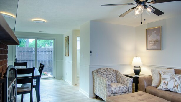 A living room with a ceiling fan, a sofa, a patterned armchair, a small table with a lamp, and a dining area with chairs and a table. - Dover Den in the Bend - Hot Tub and Pet Friendly! (South Bend)
