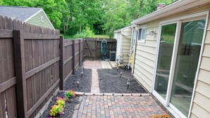 A backyard with a brick pathway, a wooden fence, and a garden bed with colorful flowers. - Dover Den in the Bend - Hot Tub and Pet Friendly! (South Bend)