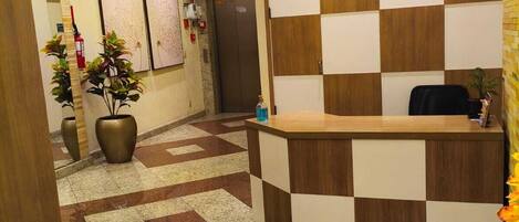 Un área de recepción con una pared a cuadros, un mostrador de recepción, un extintor y plantas en macetas.