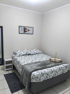 Une chambre à coucher avec un lit, une petite table de chevet blanche, un tapis gris, une œuvre d’art abstraite encadrée et un panier en osier.