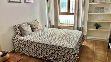 Een slaapkamer met een bed met een decoratieve spreideken, een nachtkastje met een mand, en een raam met gordijnen.
