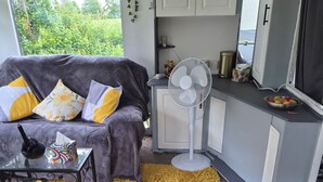 A compact living space with a sofa, a small table, a fan, and a kitchen area. - EXCLUSIVE MOBILE HOME
BASED IN COUNTRYSIDE BUT CLOSE TO AIRPORT AND TOWNS (Aldergrove)