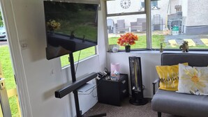 A room with a TV, a sofa, a small table, a window with a view of the outside, and a door. - EXCLUSIVE MOBILE HOME
BASED IN COUNTRYSIDE BUT CLOSE TO AIRPORT AND TOWNS (Aldergrove)