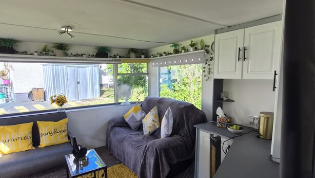 A compact living space with a sofa, a small table, a kitchenette, and a window with a view of greenery. - EXCLUSIVE MOBILE HOME
BASED IN COUNTRYSIDE BUT CLOSE TO AIRPORT AND TOWNS (Aldergrove)