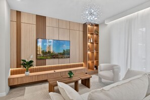 Ein modernes Wohnzimmer mit einem an einer Holzvertäfelung befestigten Fernseher, einer Bank, einem Couchtisch, einem weißen Sessel und einer dekorativen Leuchte.