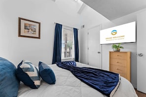 Ein Schlafzimmer mit Bett, Kissen, einem Fenster mit Vorhängen, einem an der Wand befestigten Fernseher und einem Holzkasten.