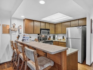 A kitchen with wooden cabinets, a stainless steel refrigerator, a microwave, and a built-in oven. There are bar stools and a granite countertop. - Giant Steps 45 Ski in/ Ski out (Brian Head)