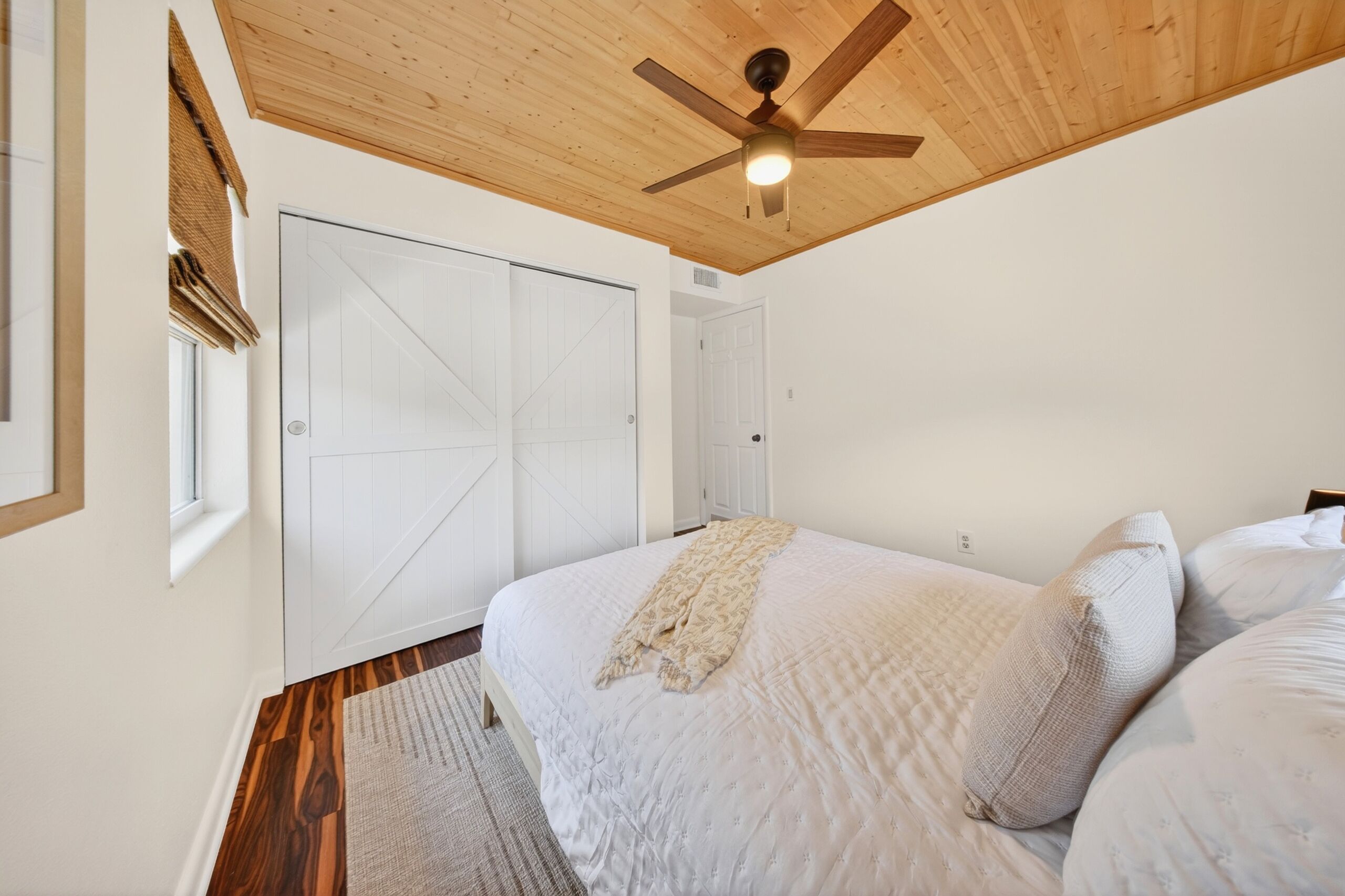 A bedroom with a wooden ceiling, a bed with a white bedspread, a ceiling fan, and a large white door.