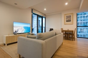 A modern living room with a flat-screen TV, a sofa, a dining table with chairs, and large windows offering a view of the city. - Bright & Modern 2BR | Pool & Wellness Facilities (Adelaide)