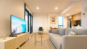 En moderne stue med et fladskærms-tv, en sofa, et lille rundt bord og et køkken i baggrunden.