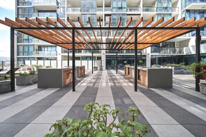 A modern outdoor terrace with a wooden pergola, seating areas, and a view of a high-rise building. - Bright & Modern 2BR | Pool & Wellness Facilities (Adelaide)