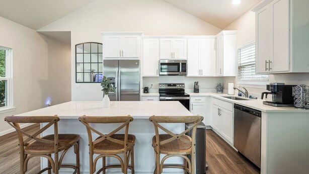 A modern kitchen with white cabinetry, stainless steel appliances, a central island with bar stools, and a view of the outdoors. - The Brainerd House · 3BR, EV Charger, Private Yard (Pensacola)