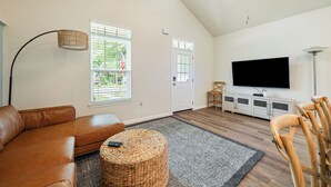 A living room with a brown leather sofa, a wicker ottoman, a television, and a white cabinet. - The Brainerd House · 3BR, EV Charger, Private Yard (Pensacola)