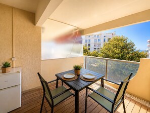 Ein Balkon mit Tisch und Stühlen, einem kleinen Kühlschrank und Blick auf Gebäude und Bäume.