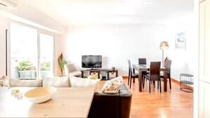 Un saló modern amb un sofà, una taula de dinar i cadires. Hi ha un televisor de pantalla plana sobre un suport, una làmpada i una taula petita amb un bol.