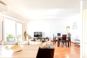 Ein modernes Wohnzimmer mit einer Couch, einem Esstisch und Stühlen. Ein Flachbildfernseher steht auf einem Schrank, zusammen mit einer Lampe und einem kleinen Tisch mit einer Schale.