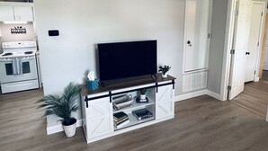 Un salon avec un téléviseur posé sur un meuble blanc, une cuisine à l’arrière-plan et une porte donnant sur une autre pièce.