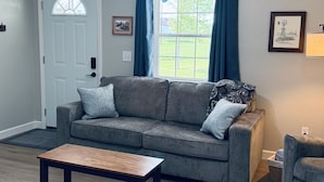Un salon avec un canapé gris, une table basse en bois, une porte blanche et une fenêtre avec des rideaux bleus.