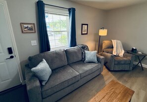 A living room with a grey sofa, a small armchair, a lamp, and a window with curtains.